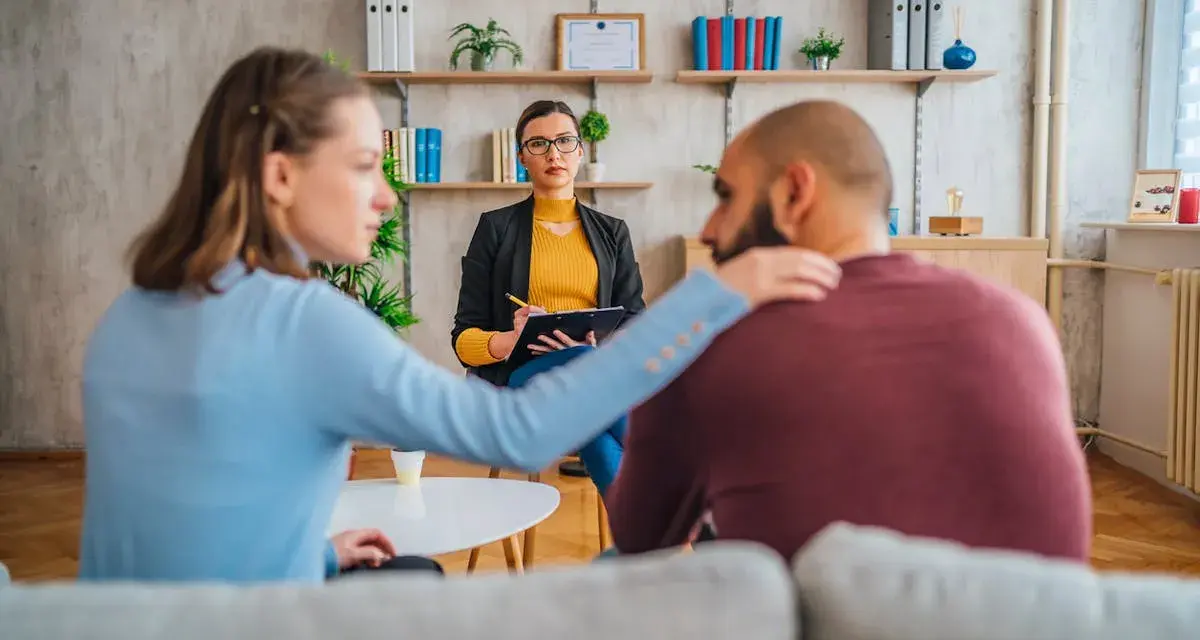 Un couple en consultation avec une psychologue conjugale. La femme pose une main réconfortante sur l'épaule de son partenaire.