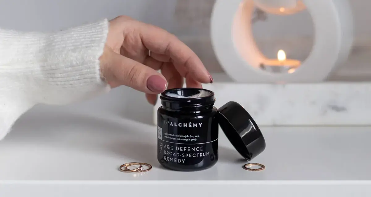 D'ALCHEMY - krem do twarzy, szyi i dekoltu. Dłoń sięga po słoiczek, w tle pali się świeca.