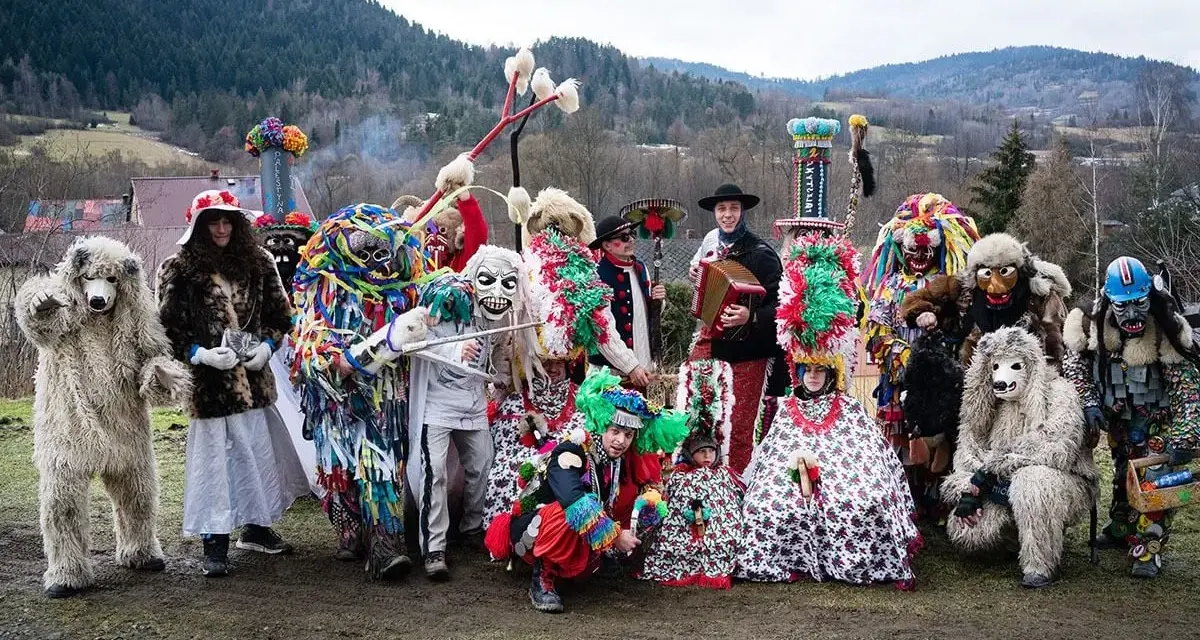 Kolorowy zwyczaj ludowy nowe latko: ludzie w fantastycznych strojach i maskach, niedźwiedzie, diabły i inne postacie pozują do zdjęcia na tle gór.