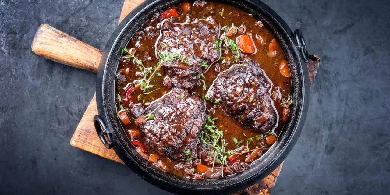 Carrilleras de ternera estofadas en salsa oscura con zanahorias y hierbas, servidas en una cazuela de hierro fundido sobre tabla de madera.