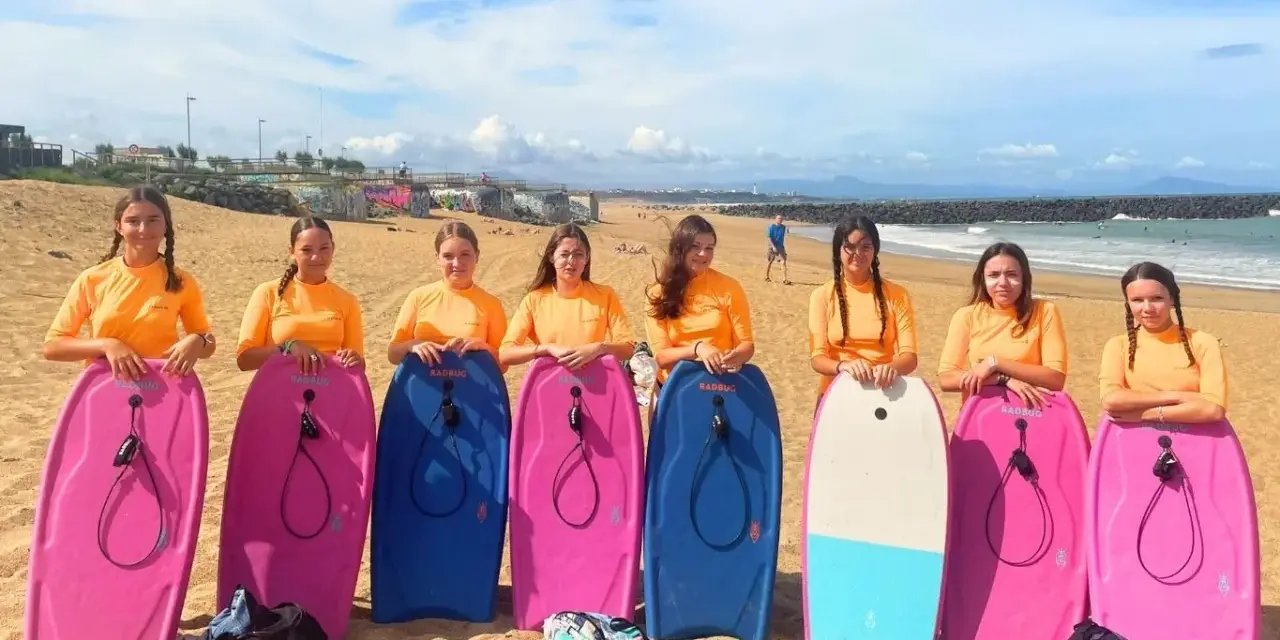 Huit jeunes filles en t-shirts orange, prêtes pour leur colonie à la mer, posent avec leurs bodyboards sur la plage.
