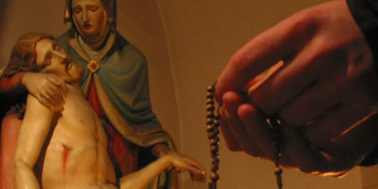 Eine Hand hält einen Rosenkranz, während die Pietà-Statue im Hintergrund zu sehen ist. Ein evangelischer Rosenkranz wird hier in Andacht gehalten.