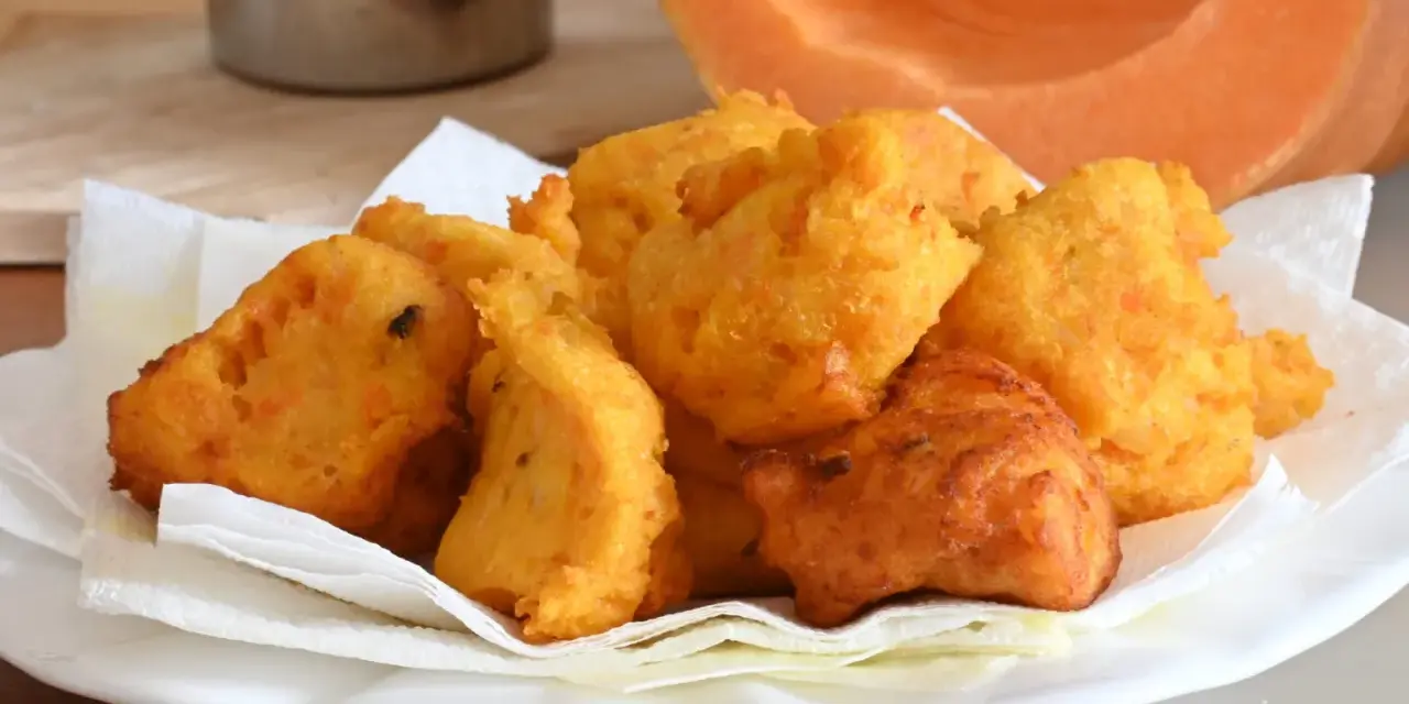 Frittelle di zucca dorate e croccanti, servite su carta assorbente, con una fetta di zucca sullo sfondo.