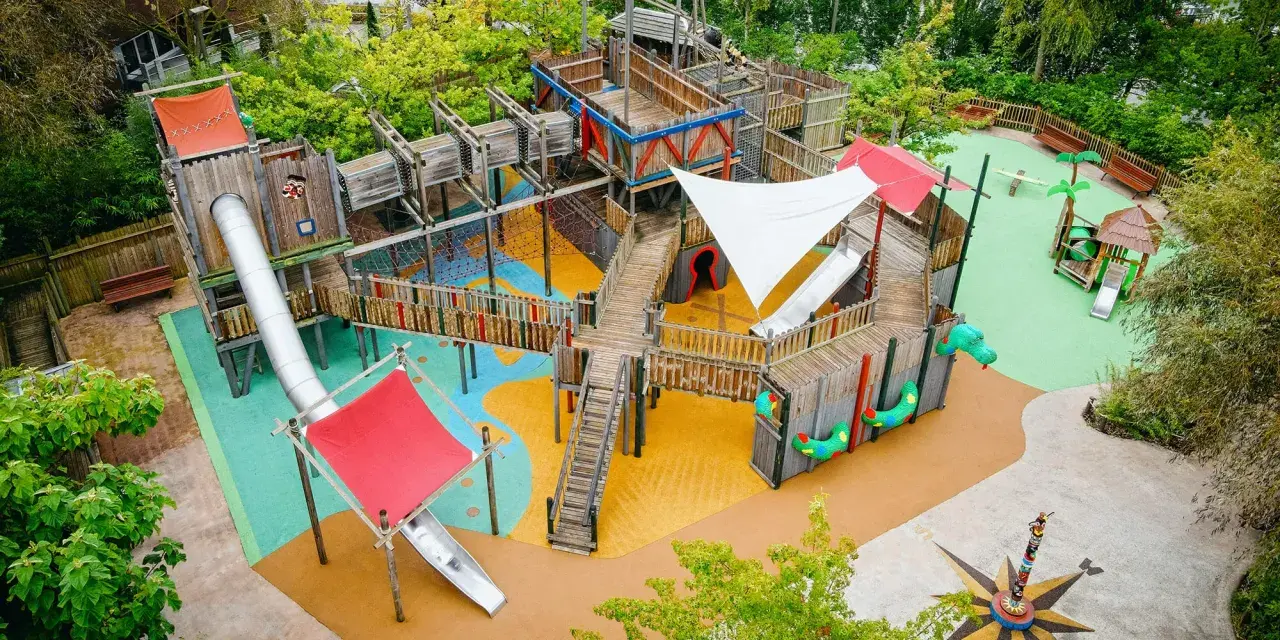 Ein riesiger, mehrstöckiger **Legoland Spielplatz** mit Rutschen, Kletternetzen und Holzbrücken, umgeben von grünen Bäumen.