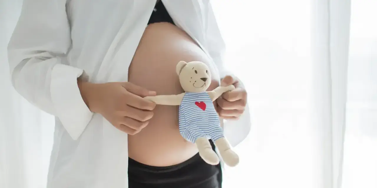 Brzuszek w 21 tygodniu ciąży, otulony białą koszulą. Dłonie trzymają misia w pasiastym ubranku, symbolizującego przyszłe macierzyństwo.
