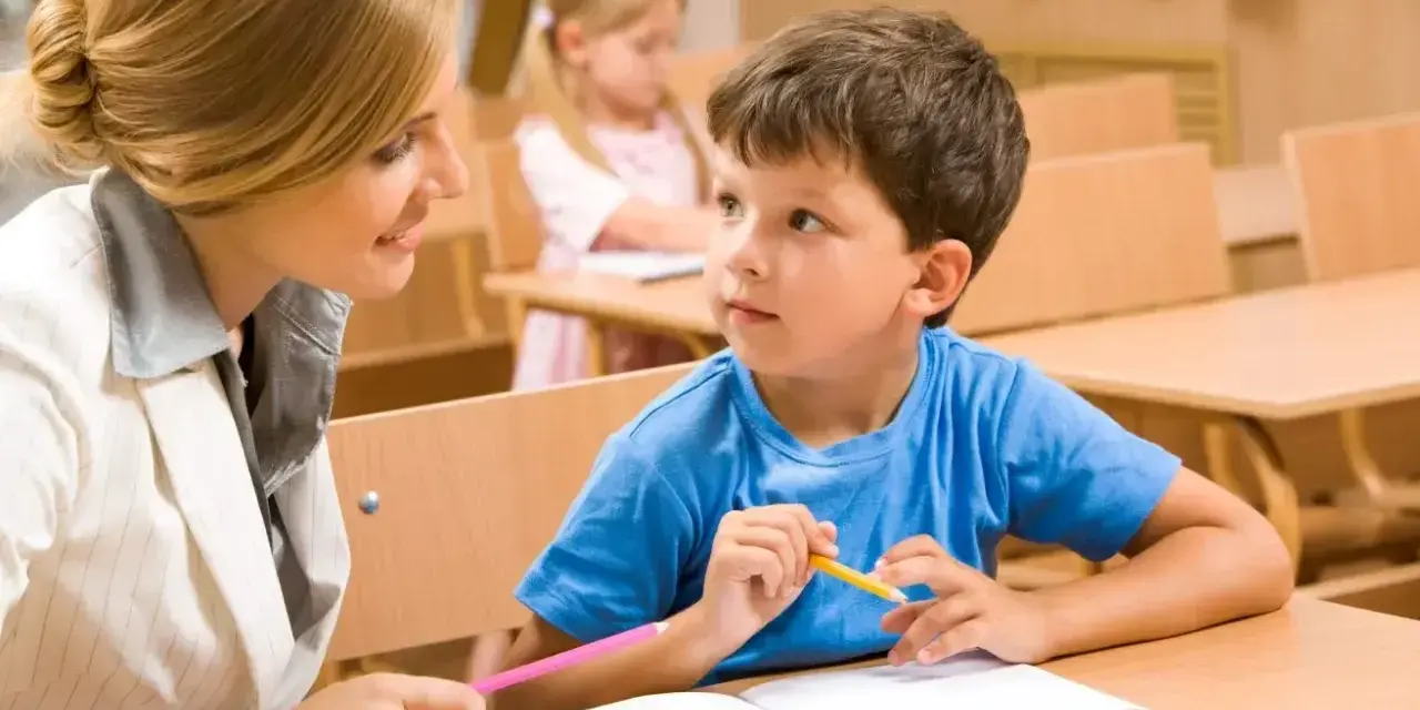 Nauczycielka pomaga uczniom w szkole. Chłopiec z zapałem słucha, trzymając kredkę.