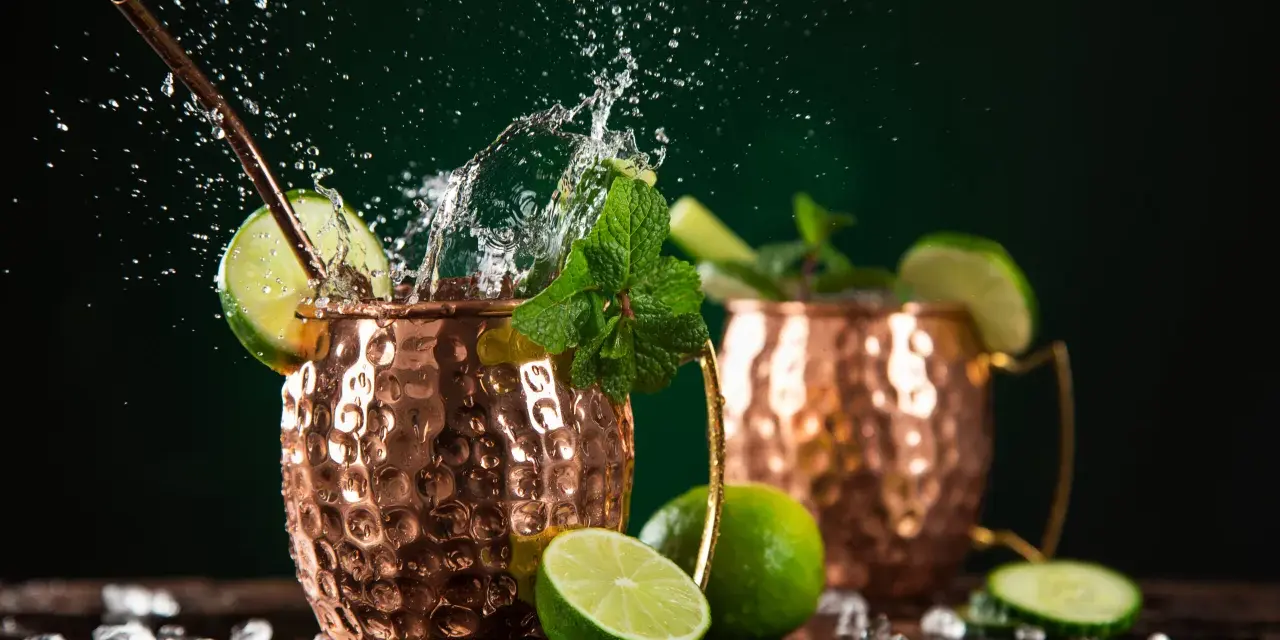 Dwa miedziane kubki z Moscow Mule i London Mule, ozdobione limonką i miętą, z bryzgami wody i lodem.