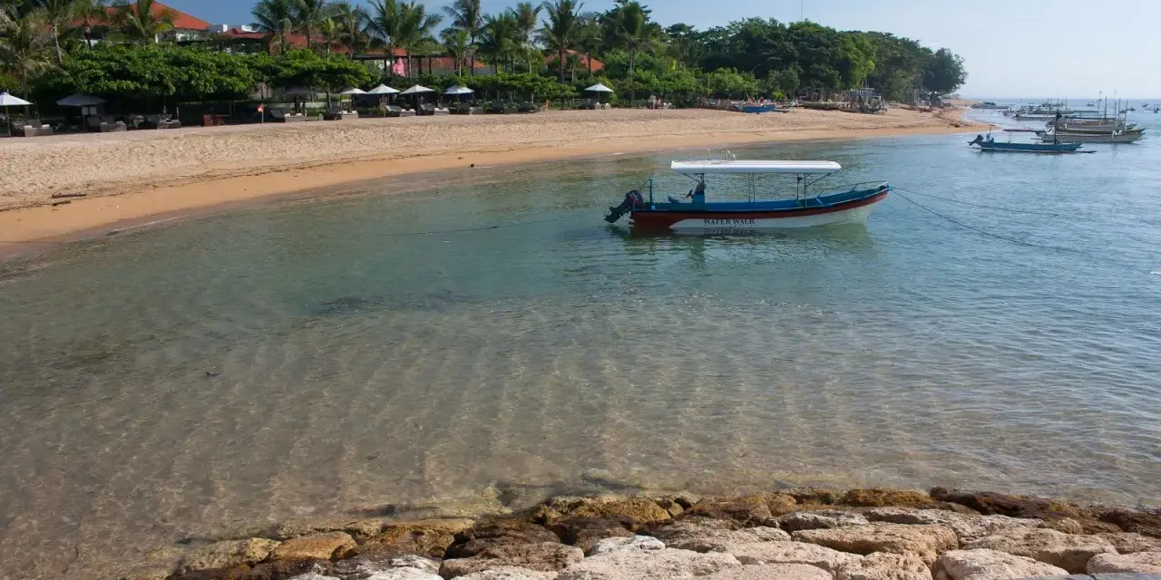 Spokojna zatoka w Sanur na Bali, z łodzią "Water Walk" kołyszącą się na turkusowej wodzie, plażą i palmami w tle.