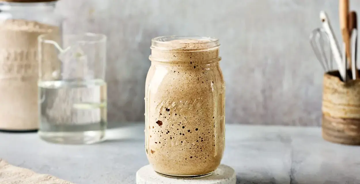 Ein Glas mit sprudelndem Sauerteig, angesetzt mit Dinkelmehl. Daneben ein Glas Wasser und ein Behälter mit Küchenutensilien.