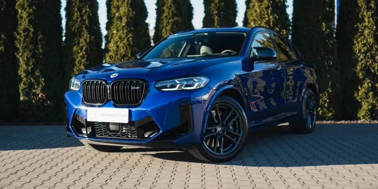 Błyszczący, niebieski suv BMW X4 M Competition zaparkowany przed żywopłotem.
