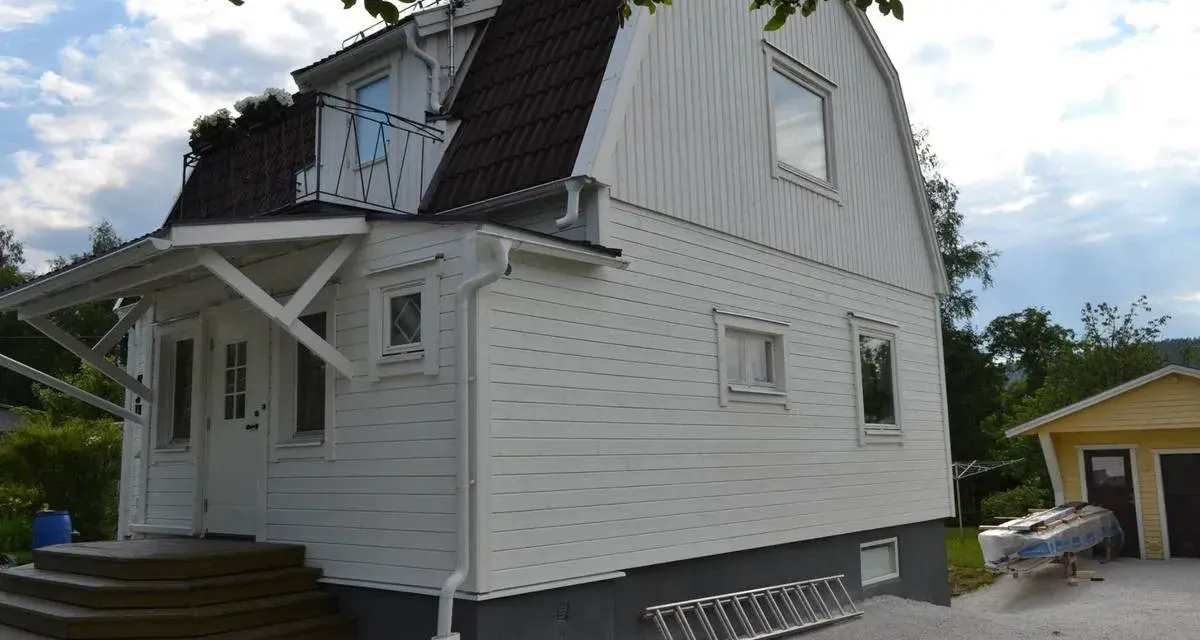 Vitt hus med ny fasad, stegar och material för byta fasad. En gul garagebyggnad syns i bakgrunden.