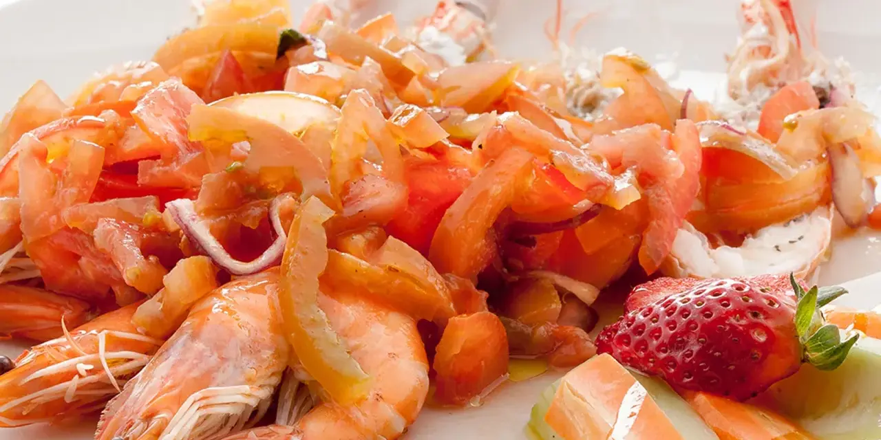 Scampi alla catalana: gamberi succulenti con pomodori freschi, cipolla rossa e una fragola decorativa. Un piatto colorato e invitante.