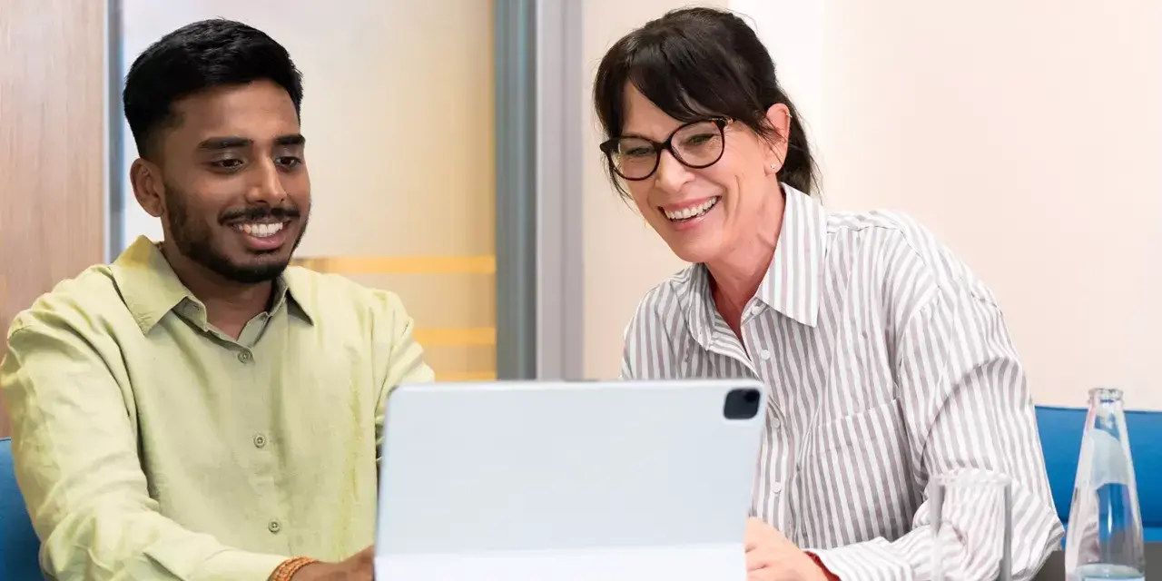 Ein junger Mann und eine Frau lachen über den Bildschirm eines Tablets. Sie sind bei einem Praktikum Studium und scheinen eine gute Zeit zu haben.