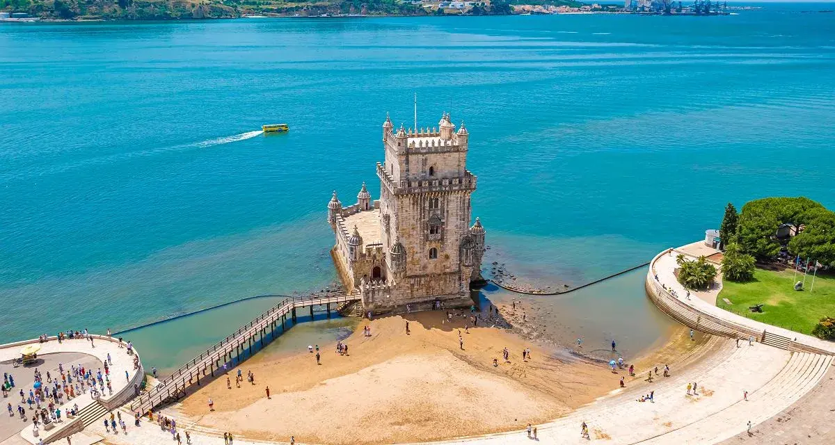 Wieża Belém, symbol kultury Portugalii, dumnie stoi nad wodą. Turyści spacerują po plaży i nabrzeżu.