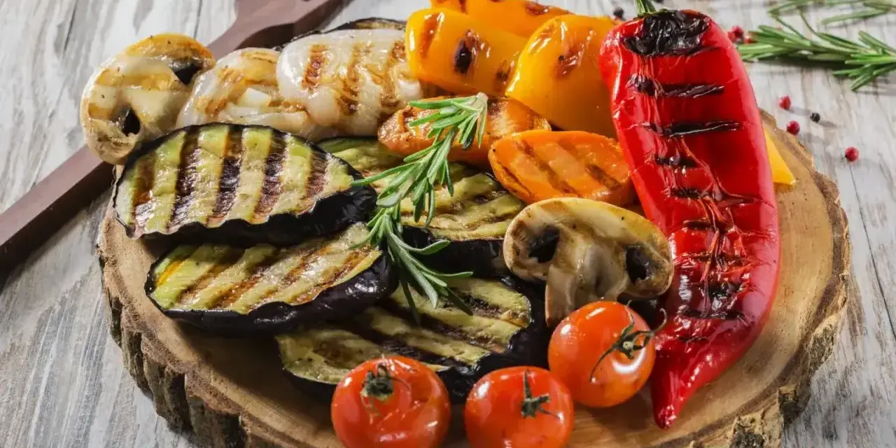 Grillowane warzywa: bakłażan, papryka, pieczarki, pomidorki i marchewka. Idealne dodatki do grilla, podane na drewnianej desce.