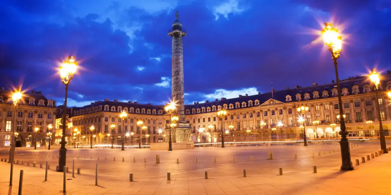 La majestuosa plaza Vendôme se ilumina bajo un cielo nocturno azul intenso, con farolas doradas y la imponente columna en el centro.