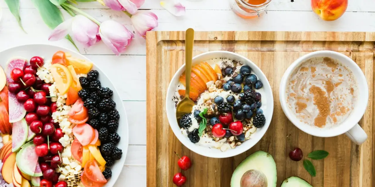 Un bol de yaourt aux fruits rouges, myrtilles et amandes, un plateau de fruits frais, un avocat et une tasse de café. Un repas sain, un aliment anti inflammatoire.