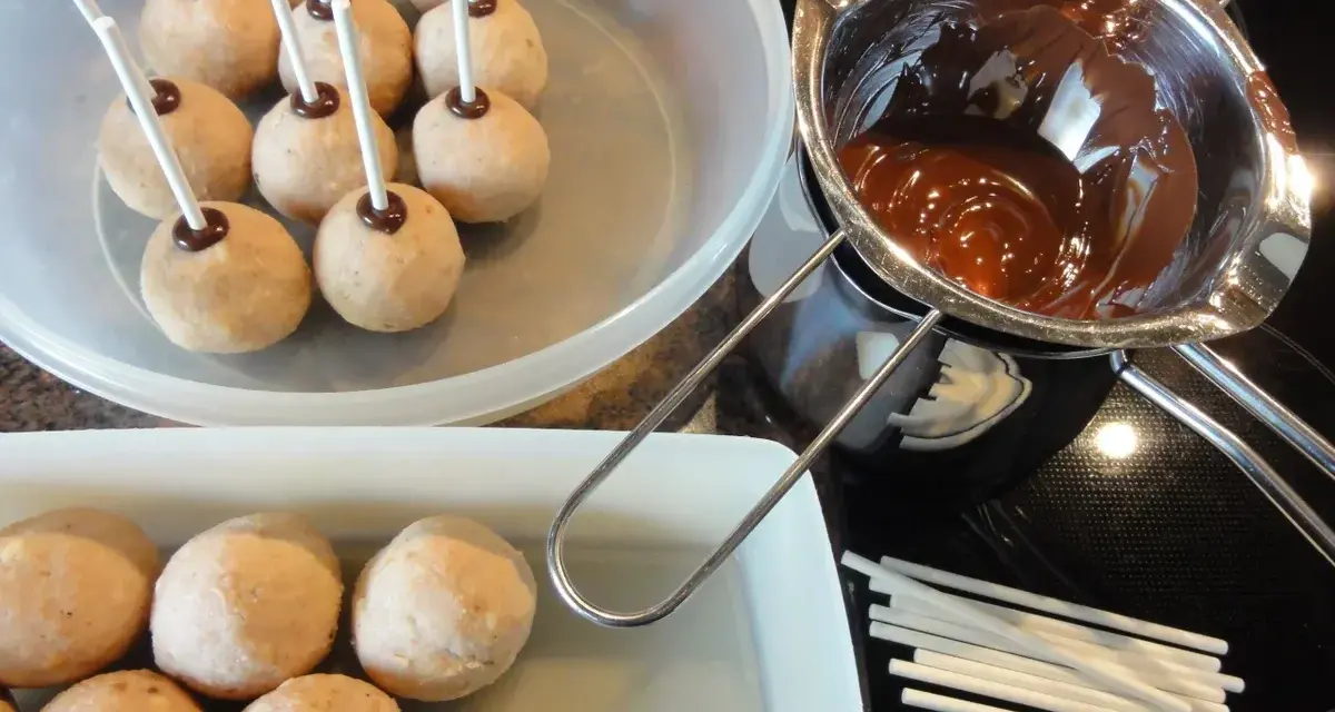 Biskuit-Cake-Pops warten auf ihre Schokoladenhülle. Geschmolzene Schokolade und Stäbchen liegen bereit.