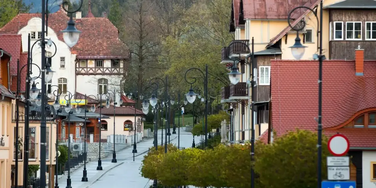 Ulica w Świeradowie Zdroju z zabytkowymi kamienicami i latarniami. Wiosenne drzewka tworzą zielony szpaler, zapraszając do odkrywania atrakcji.