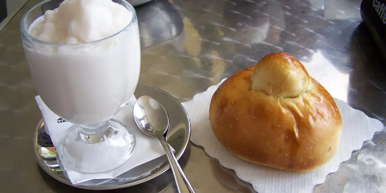 Un bicchiere di granita bianca e un soffice brioche col tuppo su un tavolo metallico.