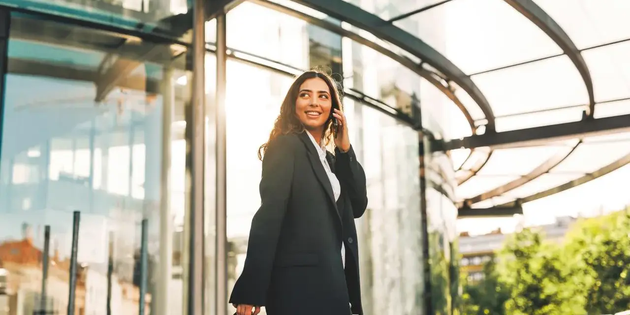 Uśmiechnięta kobieta w eleganckim stroju rozmawia przez telefon, niosąc torbę. Jej podróż po Wielkiej Brytanii trwa, a słońce odbija się w szklanej fasadzie.