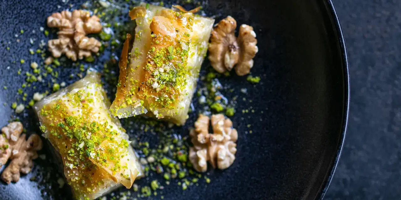 Zwei Stück Baklava, bestreut mit gehackten Pistazien und Nüssen, auf einem dunklen Teller. Ein wahrhaftiges Baklava Rezept Original.
