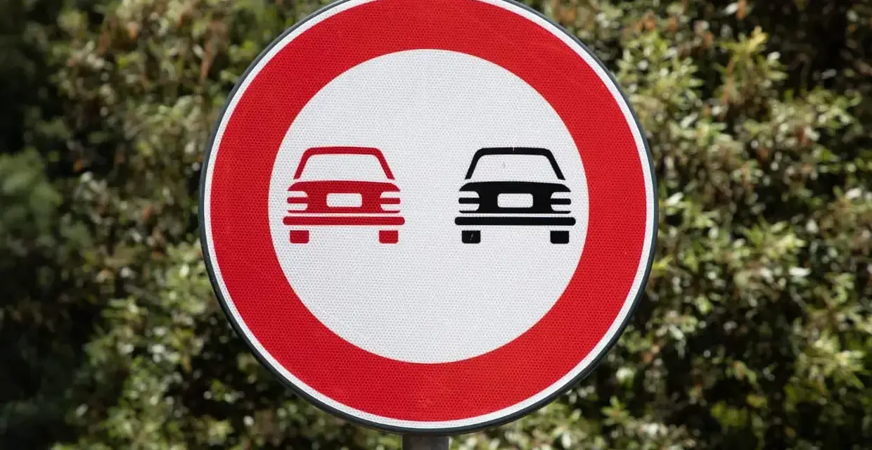 Segnale stradale art. 148: divieto di sorpasso per autoveicoli. Due auto stilizzate, una rossa e una nera, su sfondo bianco e bordo rosso.