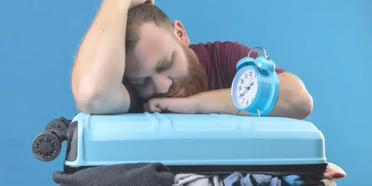 Zmęczony podróżnik śpi na walizce obok budzika. Czy to jetlag?