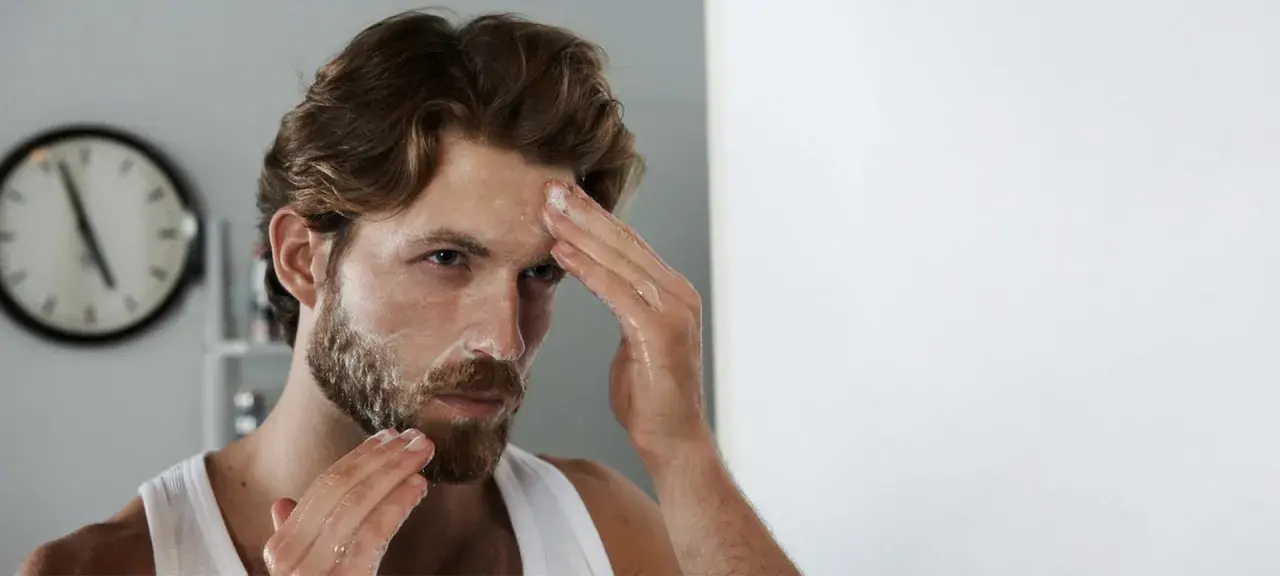 Mann mit unreiner Haut reinigt sein Gesicht mit Schaum. Er blickt in den Spiegel, während er seine Stirn einreibt.