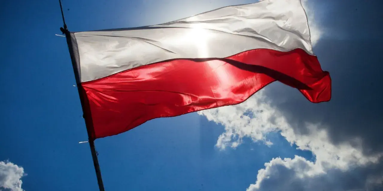 Biało-czerwona flaga Polski powiewa na tle błękitnego nieba z chmurami. Słońce przebija się przez materiał, podkreślając jego fakturę. Wymiary flagi polski są standardowe, proporcje zachowane.