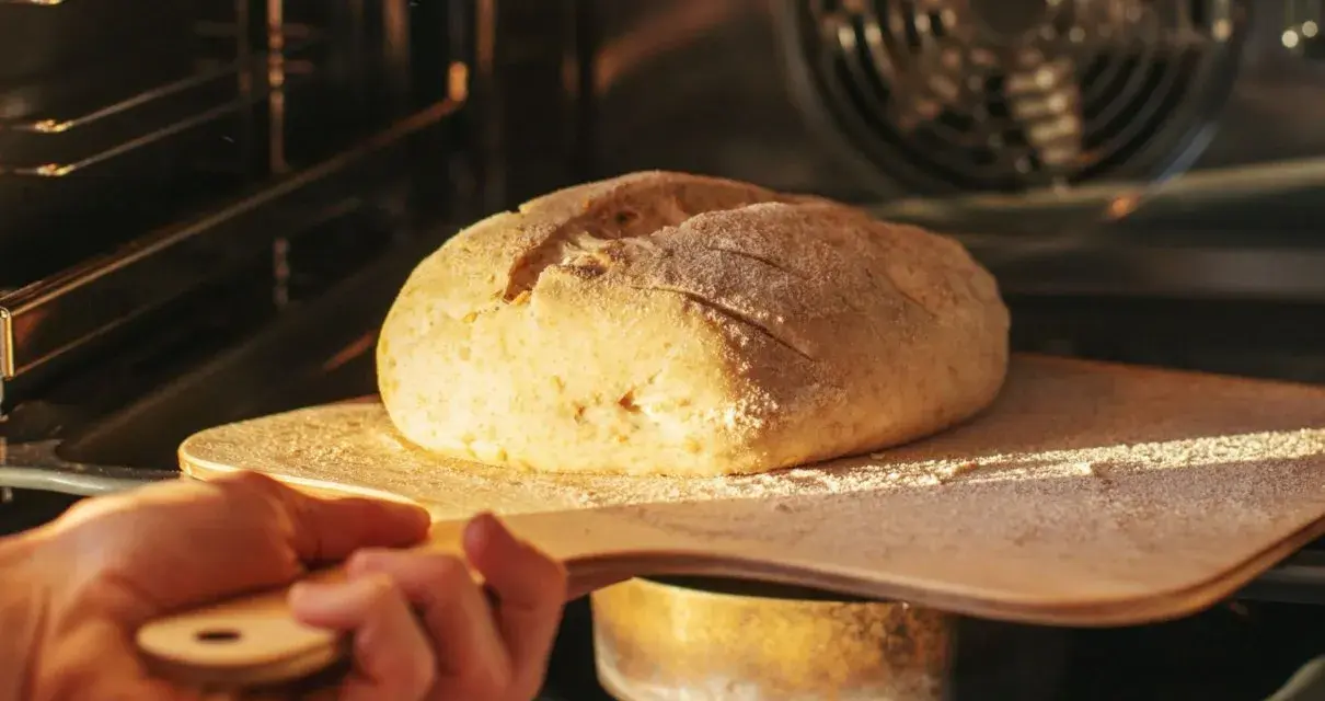 Ręka wsuwa drewnianą łopatą bochenek chleba do rozgrzanego piekarnika. To kulminacyjny moment wyrabiania ciasta na chleb.