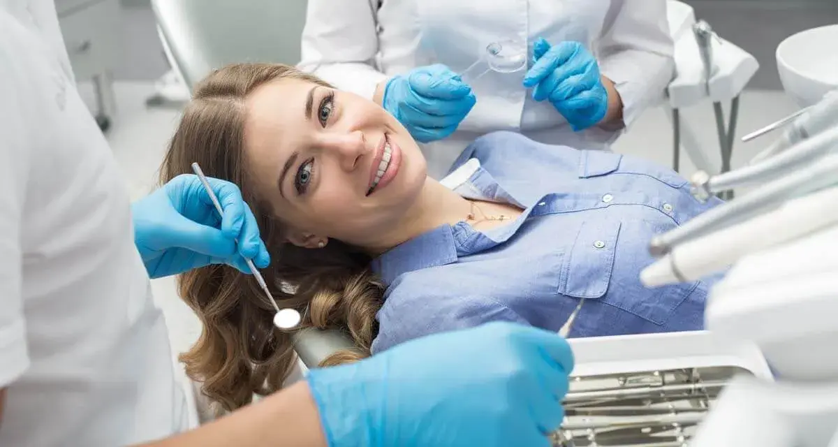 Uśmiechnięta pacjentka na fotelu dentystycznym, mimo że trochę się dentysty boi. Asystentka przygotowuje narzędzia.
