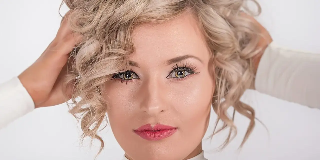 Blondynka z kręconymi włosami, z nonszalanckim gestem poprawiająca fryzurę. Jej oczy przyciągają uwagę, a usta są pomalowane na różowo.