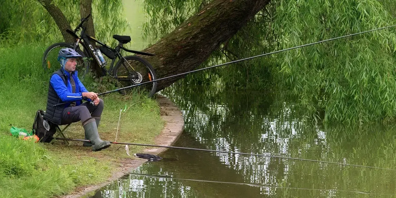 Wędkarstwo spławikowe na stawie. Mężczyzna w kasku i kamizelce siedzi na krześle, trzymając wędkę. Obok rower.