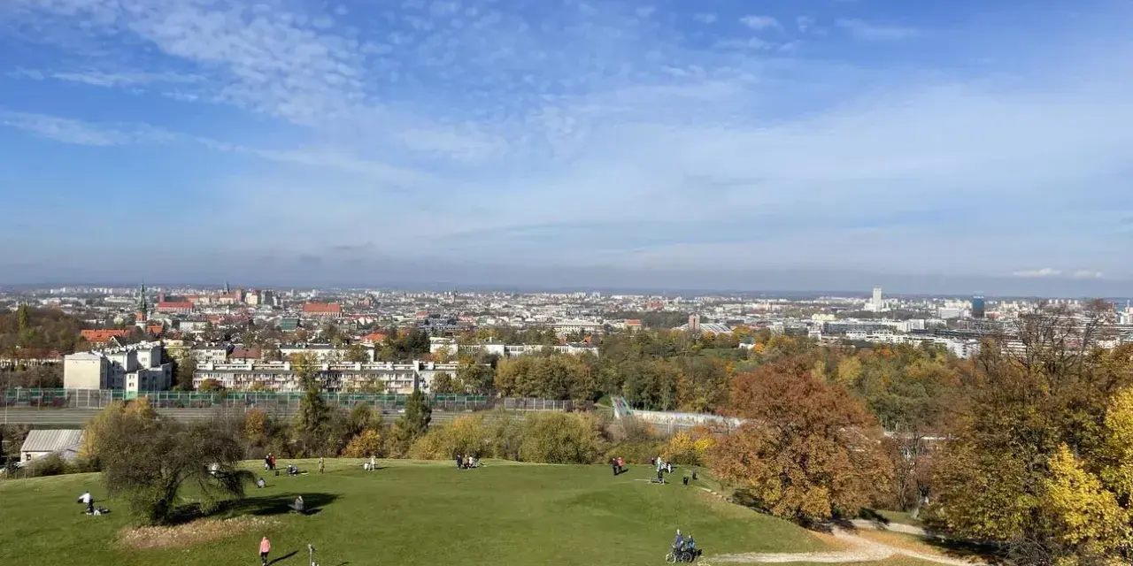Jesienny widok na Kraków z Kopca Kraka. Ludzie odpoczywają na trawie, podziwiając panoramę miasta i kolorowe drzewa.
