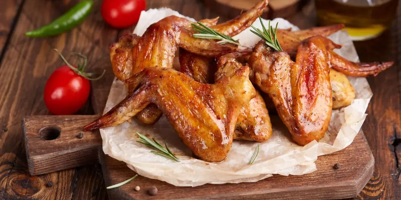 Alette di pollo dorate e succulente, cotte alla perfezione nella friggitrice ad aria, servite su carta forno con rosmarino, pomodorini e peperoncino.