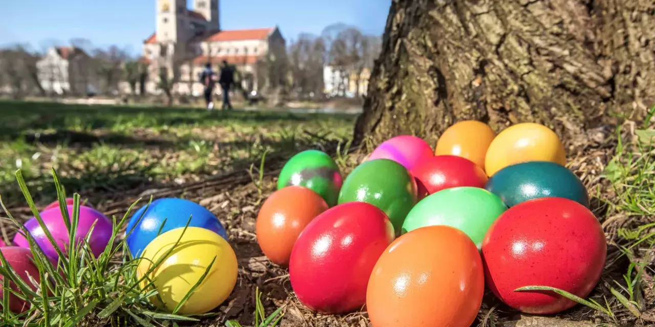 Bunte Ostereier liegen im Gras vor einem Baum. Im Hintergrund ist eine Kirche zu sehen. Die Suche nach den Eiern ist ein schöner Teil der Ostern Bedeutung.