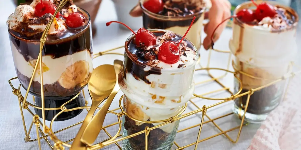 Schokoladige Dessert-Rezepte in Gläsern mit Bananen, Sahne und Kirschen. Goldene Löffel liegen bereit.