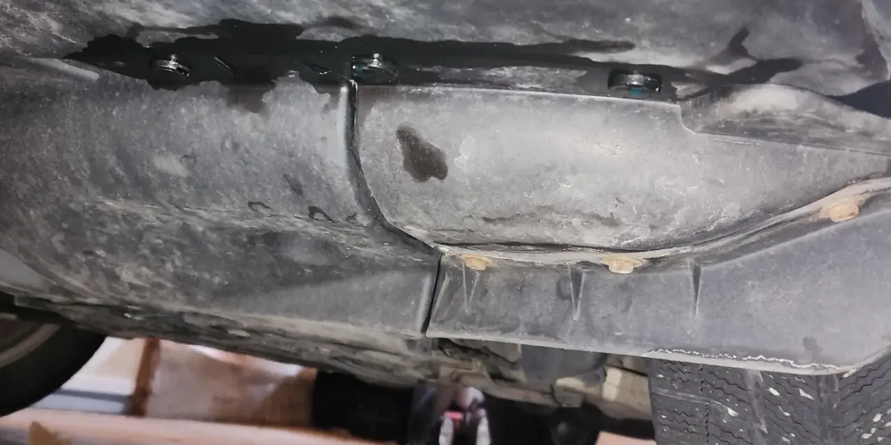 Underneath a car, dark fluid is leaking from a seam. A small puddle of blue fluid is visible, suggesting a coolant leak.