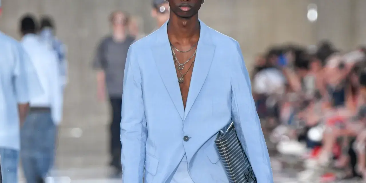 Modello con outfit uomo elegante senza giacca, completo azzurro, occhiali da sole blu e borsa a righe.