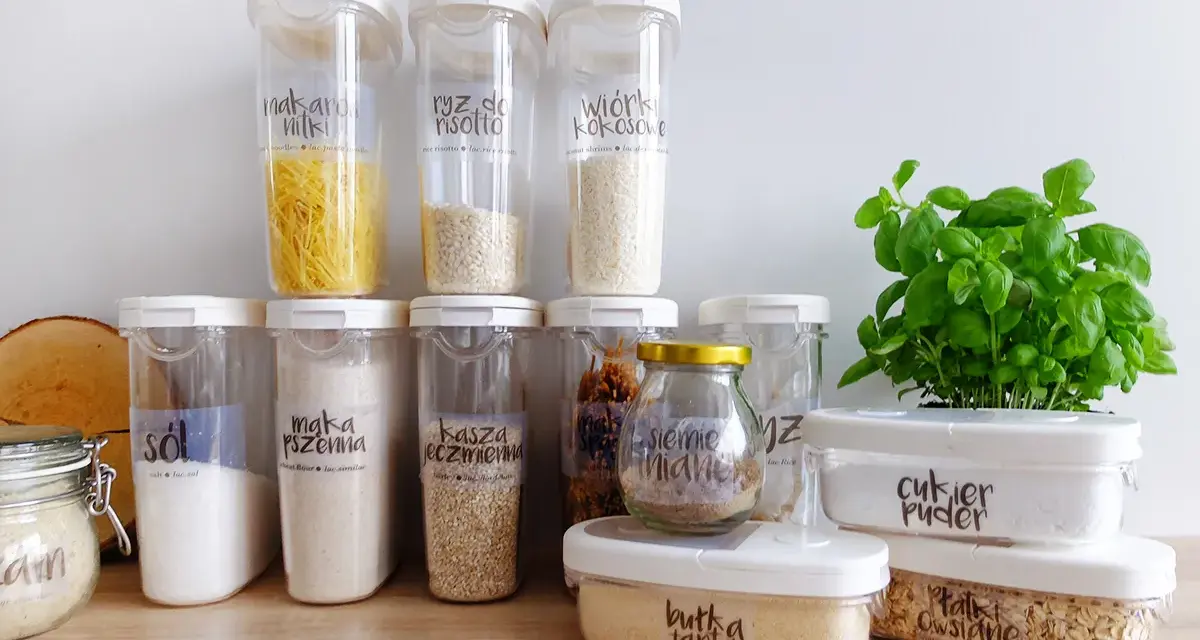 Pojemniki na żywność z etykietami pokazują, jak przechowywać pokrywki i produkty sypkie. Widać makaron, ryż, sól, mąkę, kaszę, cukier puder i płatki owsiane.