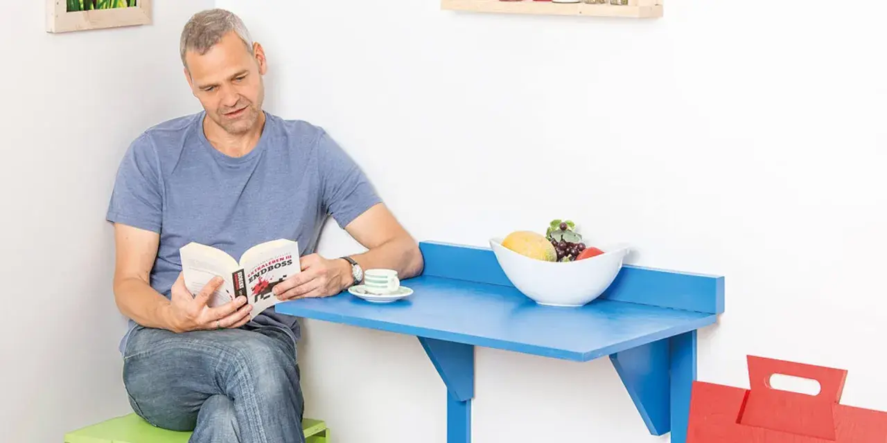 Mann liest Buch an selbstgebautem Klapptisch. Obstschale und Kaffeetasse stehen bereit.