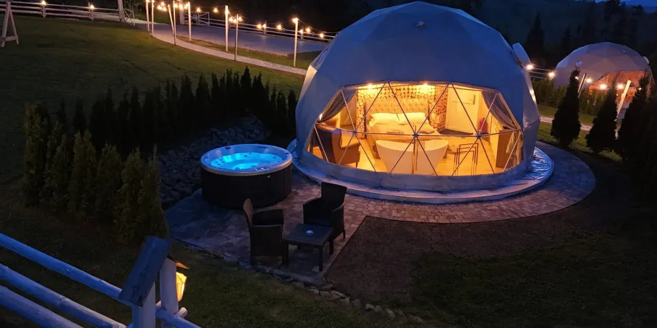 Nocny widok na luksusowy camping w Tatrach. Dwa kopułowe namioty, jeden z wanną i łóżkiem, drugi z oświetleniem. Obok jacuzzi i meble ogrodowe.
