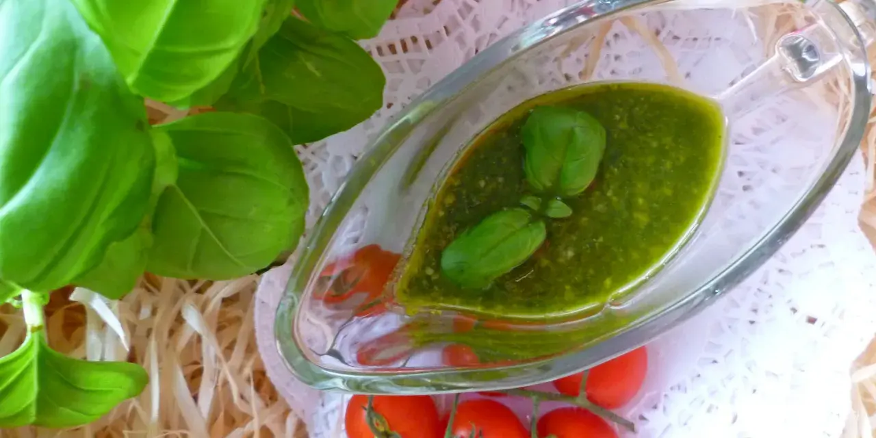 Aromatyczne pesto bazyliowe w szklanej sosjerce, ozdobione listkami bazylii, obok gałązki pomidorków koktajlowych.