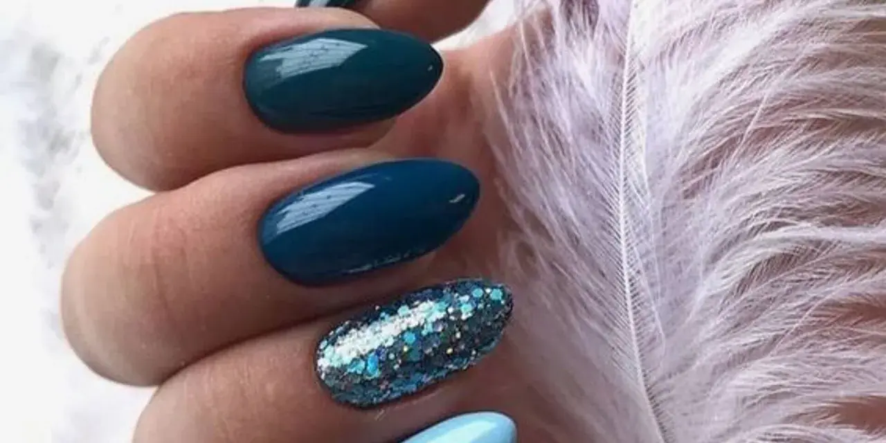 Unghie per l'inverno: unghie blu petrolio e azzurre con glitter scintillanti, accanto a una soffice piuma bianca.