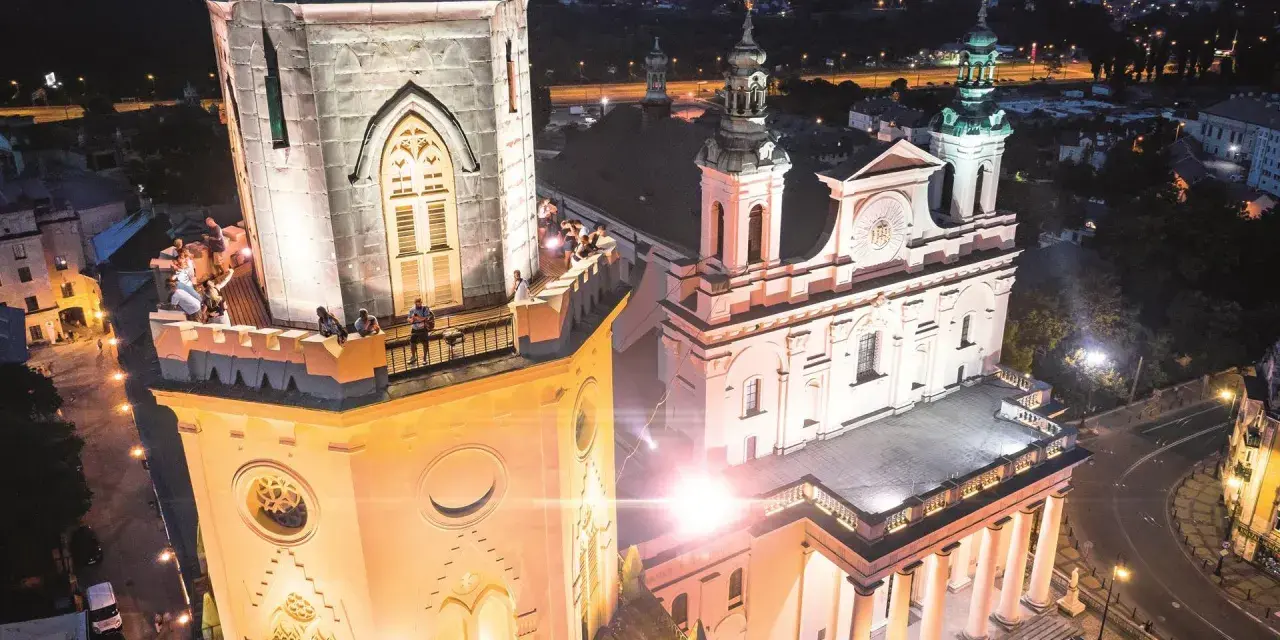 Wieża trynitarska nocą, z ludźmi na balkonie i oświetloną fasadą kościoła.