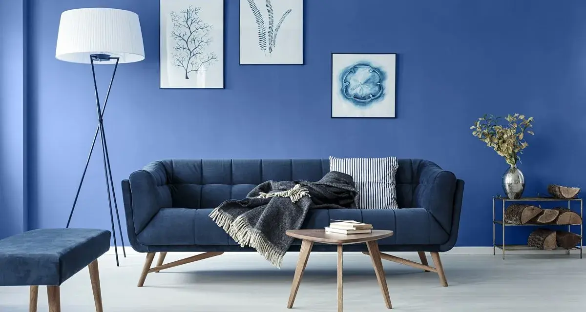 Niebieska sofa z szarym kocem i książkami na stoliku. Na ścianie obrazy z motywami roślinnymi i morskim.