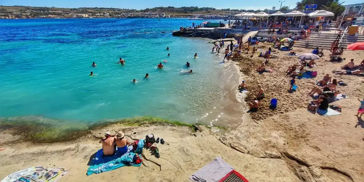 Słoneczna plaża na Malcie w lutym. Ludzie kąpią się w turkusowej wodzie, opalają na piasku i odpoczywają pod parasolami.