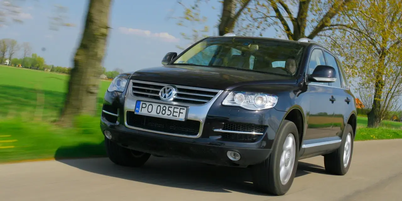 Czarny VW Touareg 3.0 TDI w ruchu, z widocznymi wadami lakieru i drobnymi niedoskonałościami, na tle zielonych pól i drzew.