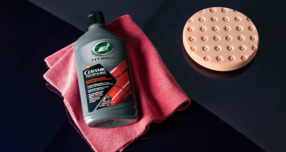 En flaska Turtle Wax Hybrid Solutions Ceramic Polish & Wax, en rosa mikrofiberduk och en polertrissa ligger på en mörkblå yta.